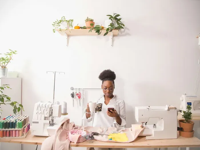 Young woman working in sewing studio using smartphone while surrounded by fabrics and sewing equipment on table